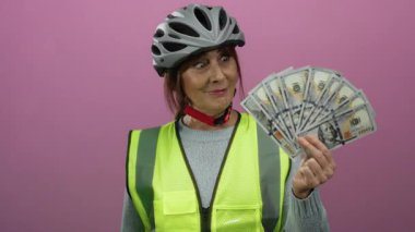 Miğfer ve yelek giyen İspanyol kadın Amerikan dolarlarını pembe duvara dayayıp finansal başarıyı güvenlikle sembolize ediyor..