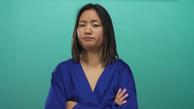 Çapraz kollu bir kadın, yeşil bir arka plana karşı mavi bir bluz giyerek poz veriyor, ifadeler ve jestlerle duygularını sergiliyor, derin düşüncelere dalmış bir ruh halini vurguluyor..