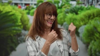 Yeşil bitki ve patikalarla çevrili bir parkta kollarını açmış gülümseyen yaşlı İspanyol kadın doğa ve mutluluğun neşeli bir sahnesini yaratıyor..