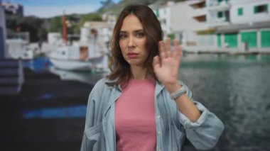 Genç İspanyol bir kadın, açık mavi gökyüzünün altında, liman kenarında rahat bir hava taşıyan, sıradan bir kot gömlek ve pembe bir bluz giyerek, deniz kenarında kameraya el sallıyor..