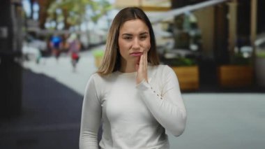 Dişleri ağrıyan genç bir kadın kalabalık bir sokakta dikiliyor, beyaz bir gömlek giyiyor ve üzgün görünüyor, yayalar arka planda bulanık bir şekilde yürürken yanağına dokunuyor..