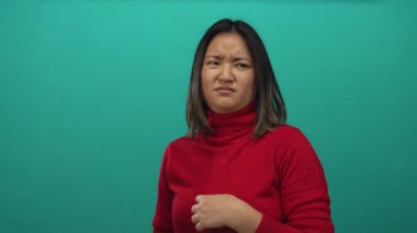 İzole edilmiş yeşil arka plandan iğrenen bir kadın, kırmızı kazak giyiyor, genç bir kadın negatif duygu gösteriyor, Asya kökenli, zümrüt zemin üzerinde duruyor..