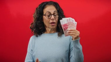 İzole edilmiş kırmızı arka planda İzlanda banknotları tutan kadın, orta yaş özellikleriyle merakını dile getiriyor ve elde bulunan paraya ilgi çekici bir bakış atıyor..