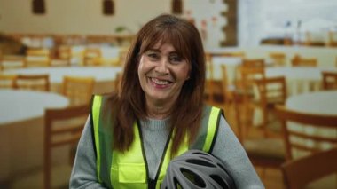 Üst düzey İspanyol kadın, kapalı bir restoranda gülümsüyor, güvenlik yeleği giyiyor ve kaskını tutuyor, ahşap sandalyeler ve masalarla çevrili, sıcaklık ve güven saçıyor..