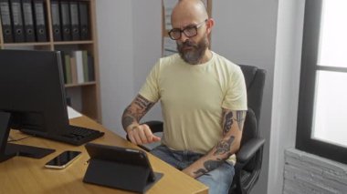 Masaüstü bilgisayarı ve akıllı cihazları olan bir ofiste esneyen sakallı ve dövmeli İspanyol bir adam.