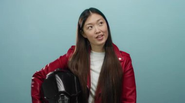 Kırmızı ceketli kadın kaskını mavi arka planda tutuyor. Düşünceli ve özgüvenli görünüyor. Stüdyo ışıklarıyla canlandırıcı bir çekiciliği var.