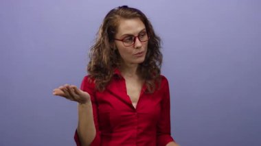 Kırmızı gözlüklü ve gömlekli, mor bir arkaplan karşısında el kol hareketi yapan kadın, belirsizliği ifade eden sakin bir yüz ifadesiyle kafa karışıklığı gösteriyor..
