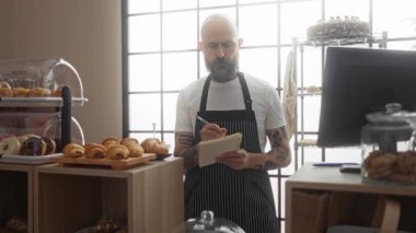 Pasta fırınında yeni pişmiş hamur işleri ve pastalar sergilenmiş, çizgili önlük giymiş, fırın eşyaları ve bilgisayarla çevrili ve kapalı bir dükkan ortamında duran bir adam not defterinde yazıyor.