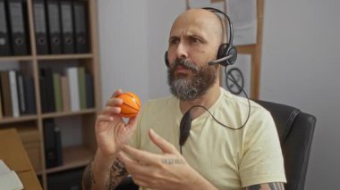 Ofiste sakallı bir adam, kitap raflarıyla çevrili bir şekilde konuşurken ve masada otururken stres topu tutuyor.