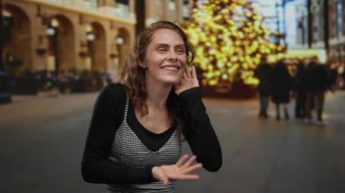 Genç bir kadın hareketli bir şehir sokağında kışın neşeli ışıklar ve bulanık kalabalıkla dinleyici jestler yapıyor..