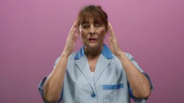 Kıdemli İspanyol kadın üniformalı temizlikçi elleri pembe arka planda şakakların üzerinde stresli görünüyor, hüsran ve yorgunluk taşıyor..