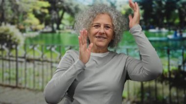 Gri saçlı bir kadın, güneşli park ortamında elleri ile, yemyeşil bir zemin üzerinde, günlük gri bir gömlek giyerek, neşe ve yaratıcılık ifade eden hareketler yapar..