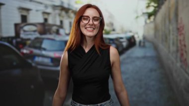 Şehir caddesinde arabaları kenarda park etmiş bir şekilde gülümseyen bir kadın, şehir ortamında neşe ve güven sergiliyor, sıradan moda ve kızıl saçlar sergiliyor..