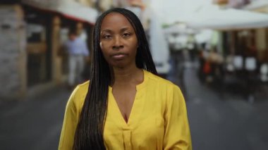 Bir kadın sokak kafesi ambiyansıyla terasta tefekkür ediyor, sarı giyiniyor, açık hava manzarası sergiliyor ve siyah saçlı..