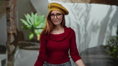 Gözlüklü ve sarı şapkalı bir kadın arka planda bitkilerle dikiliyor. Rahat ve kendine güvenen kırmızı elbiseli bir kadın..
