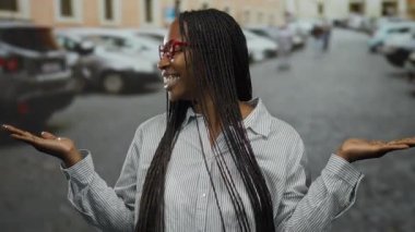 Kıvırcık saçlı, gözlüklü bir kadın şehir arka planında, park etmiş arabalarla neşeyle gülümsüyor, canlı şehir hayatı ve dinamik açık hava atmosferi sergiliyor..