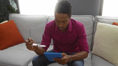 Genç adam evdeki modern yaşam tarzını ve teknolojisini sergilemek için rahat oturma odasında tablet kullanırken gözlükleri ayarlıyor..