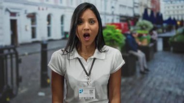 Kalabalık bir caddede, dışarıda duran genç bir kadın. Şaşırmış bir ifadeyle, beyaz bir gömlek ve kimlik kartı takıyor. Şehirde çeşitli duygular sergiliyor..