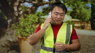 Akıllı telefondan bağıran Asyalı adam parkta güvenlik yeleği giyiyor. Arka planda ağaçlar ve bitkiler var..