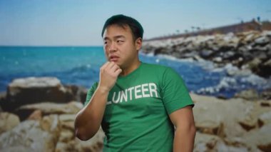 Deniz kenarında yeşil tişörtlü ve gönüllü mesajlı genç bir adam. Parlak güneş ışığında plaj manzarasının tadını çıkarıyor.