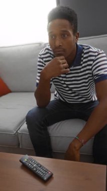 Oturma odasındaki bir kanepede oturan genç adam, düşünceli ve düşünceli görünüyor, masada uzaktan kumandayla..