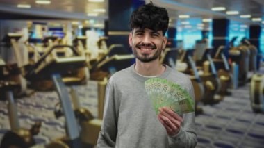 Spor salonunda spor aletleriyle Rus rublesi taşıyan bir adam, kapalı bir spor salonunda güvenle gülümsüyor..