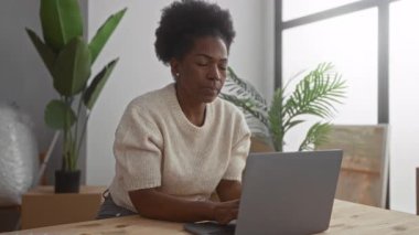 Modern oturma odasında dizüstü bilgisayar kullanırken gülümseyen bir kadın. Etrafı bitkiler ve kutular ile çevrili. Rahatlamış ve rahat bir atmosferi olan yeni bir ev ortamı öneriyor..