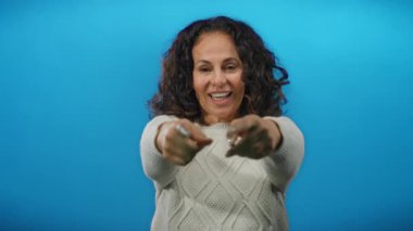 Gülümseyen bir kadın açık açık parlak mavi arkaplan karşısında neşeli yüz ifadesini ve neşeli, izole bir ortamda davetkar hareketlerini vurgular..