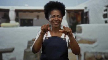 Önlüklü Afro-Amerikalı bir kadın, şehir dışında bir arka plana karşı hoş geldin jesti yaparak, bir şehir caddesinde güvenle gülümsüyor..