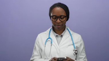 Akıllı telefondan mesaj atan kadın doktor. Mor duvar arkasında steteskop ve gözlüklerle izole edilmiş. Tıp alanında iletişim ve teknoloji olduğunu gösteriyor..