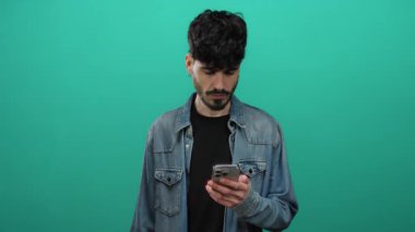 Kot ceketli genç adam, izole edilmiş yeşil arka plana karşı akıllı telefon kullanıyor. Cihazıyla etkileşime girerken meşgul ve anlamlı görünüyor..