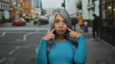 Yoğun bir şehir sokağında, arka planda arabaları ve yayaları olan sokak hayatını gözler önüne seren, neşeli bir ifade sergileyen gri saçlı son sınıf kadını..