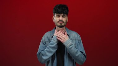 Kırmızı arka planda izole edilmiş genç adam üzgün görünüyor. Ellerini boğazına koyuyor. Kot ceket giyiyor.