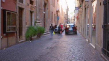 Roma 'da bir kaldırım taşı caddesinde yürüyen insanların bulanık görüntüsü. Parlak güneş ışığı altında arka planda uzanan eski binalar kentsel yaşamı ve mimariyi gözler önüne seriyor..