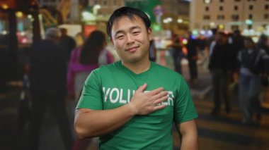 Yeşil gönüllü gömlekli genç adam, şehir sokaklarında geceleri gülümsüyor, şehir ışıkları ve bulanık yayalarla çevrili, toplum ruhunu somutlaştırıyor..