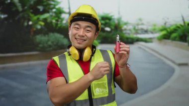 İnşaat işçisi, şehir ortamında elinde bir tüple dışarıda gülümsüyor, güvenlik giysileri giyiyor. Sıkı bir şapka ve yüksek görüş mesafesi olan bir yelek, odak noktası olarak e-sigara sergiliyor..