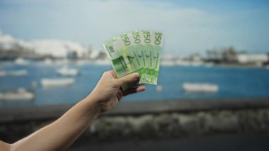 Norveç kronu taşıyan bir adam, arka planda tekne ve su bulunan bir sahil limanında açık hava ve deniz yaşam tarzını ön plana çıkarıyor..