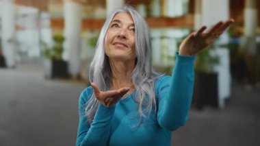 Gülümseyen kadın, şehir caddesinde elini uzatıyor. Gri saçlı, parlak mavi kazaklı, açık havada mutlu bir atmosfer yaratıyor.