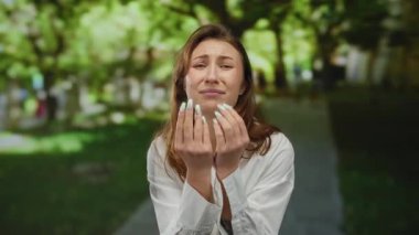 Neşeli yüz ifadeleri olan genç bir kadın, yeşil ve ağaçlarla çevrili güneşli bir parkta parmaklarını çimdikleyerek komik bir an yaşatıyor..