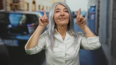 Yaşlı bir kadın, kır saçlı, sokaklarda sevinçle parmaklarını kaldırıyor. Sanki şehir manzarasının üzerinde ilham verici bir şey varmış gibi..