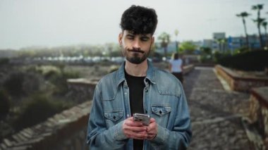 Kentsel sokaktaki genç İspanyol adam şehir manzarasının arka planıyla akıllı telefonu karşılaştırırken dalgın görünüyor..