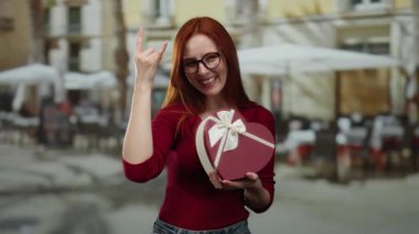 Terasta hediyeyle gülümseyen, gözlüklü ve kırmızı gömlekli bir kadın şehir restoranında neşe saçıyor..