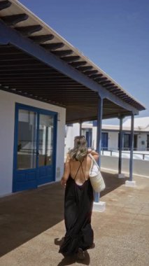 Lanzarote kanarya adalarında güneşli bir günün tadını çıkaran bir kadın siyah bir elbise giyer ve arkasında mavi kapılar ve beyaz duvarlar olan gölgeli bir verandanın altında duran bir kamış çanta taşır.