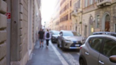 Roma 'da insanların yürüdüğü ve eski dar bir yol boyunca park etmiş arabaların parlak güneş ışığı altında tarihi bir mimari sergilediği bulanık bir sokak manzarası..