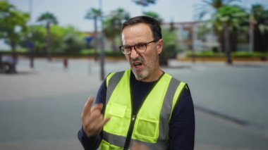 Güvenli yelek giyen bir adam şehir parkında dışarıda taş el hareketleri yapıyor, heyecanını dışa vuruyor, şehir geçmişi bulanık..