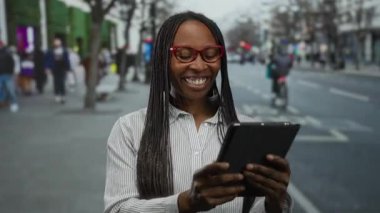 Örgü örgülü ve gözlüklü kadın şehir merkezinde tablet kullanıyor. Arka planda insanlar var. Şehir yaşamını ve teknolojiyi yansıtıyor..