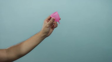 Özel bakım ürünlerinde çevre dostu tercihi ve sağlık bilincini vurgulamak için pembe regl bardağını mavi arka planda tutan adam.