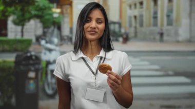 Kadın parmaklarıyla küçük bir jest yapıyor, şehir sokağında kurabiye tutuyor, beyaz gömlek ve rozet giyiyor, açık hava ortamında en küçük boyut fikrini iletiyor..