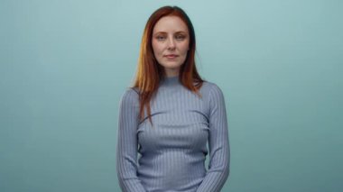 Mavi arka planda, göğsünde bir elle duran, gri bir kazak giyen, kızıl saçlı, yetişkin ve güzel bir yüz ifadesi olan, sakin ve sakin bir atmosferi olan bir kadın..