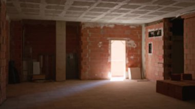 Tamamlanmamış, kırmızı tuğlalı duvarları ve beton tavanı olan boş bir oda. İnşaat alanındaki bir geçitten gelen doğal ışıkla aydınlatılmış.